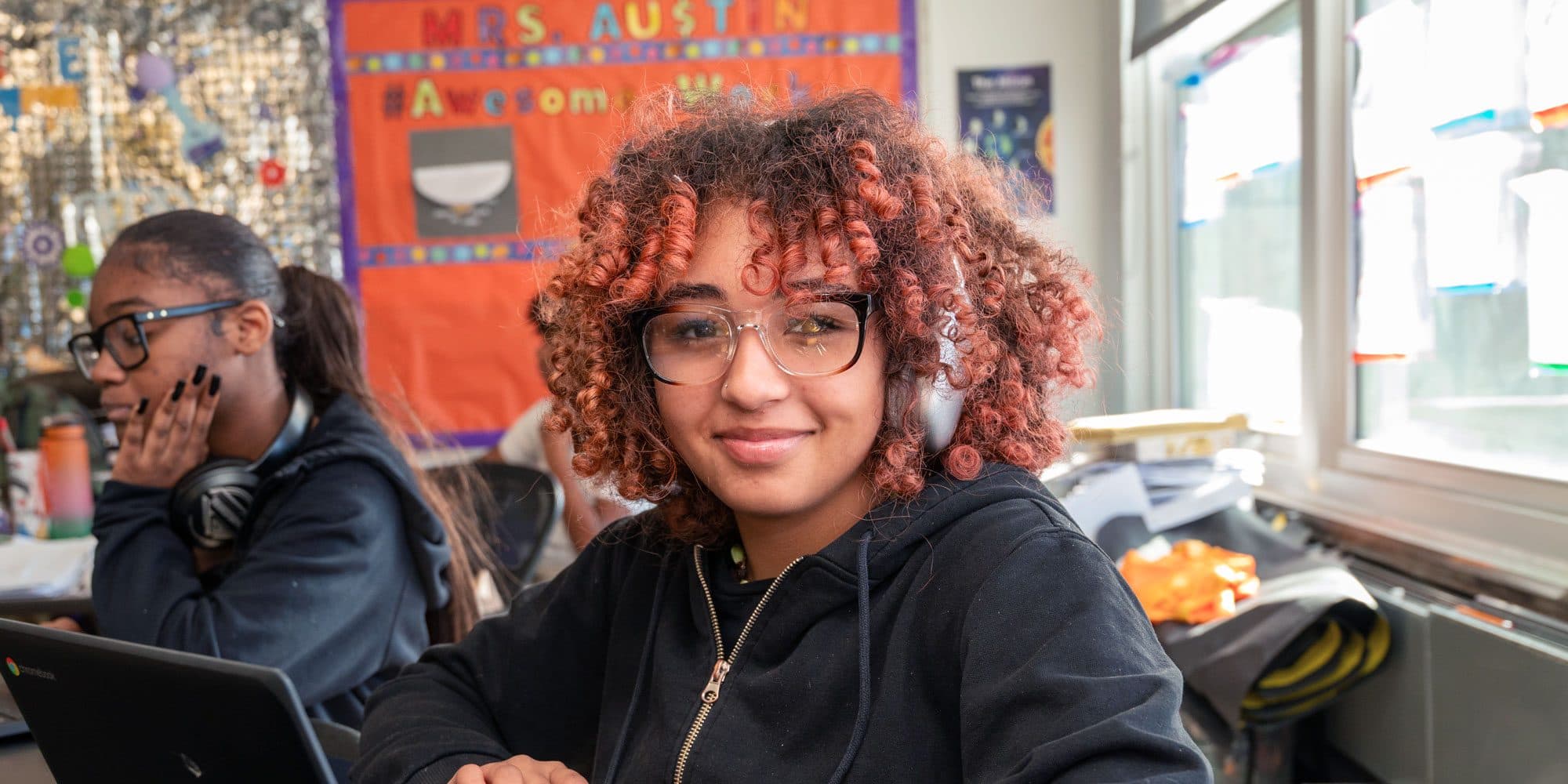 Smiling student with glasses and headphones in a classroom.