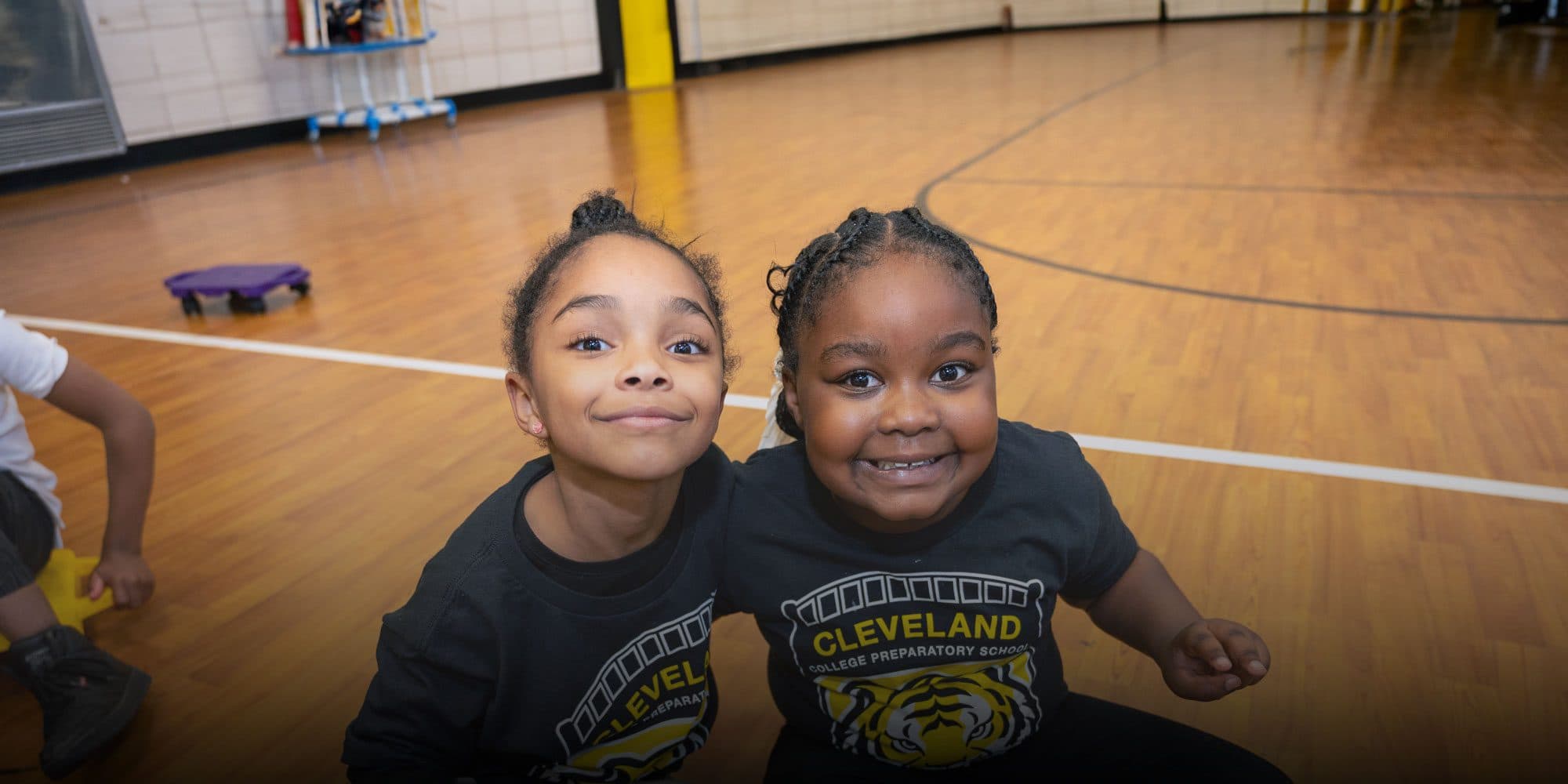 Two smiling Cleveland College Prep students in school shirts sitting together in the gym.