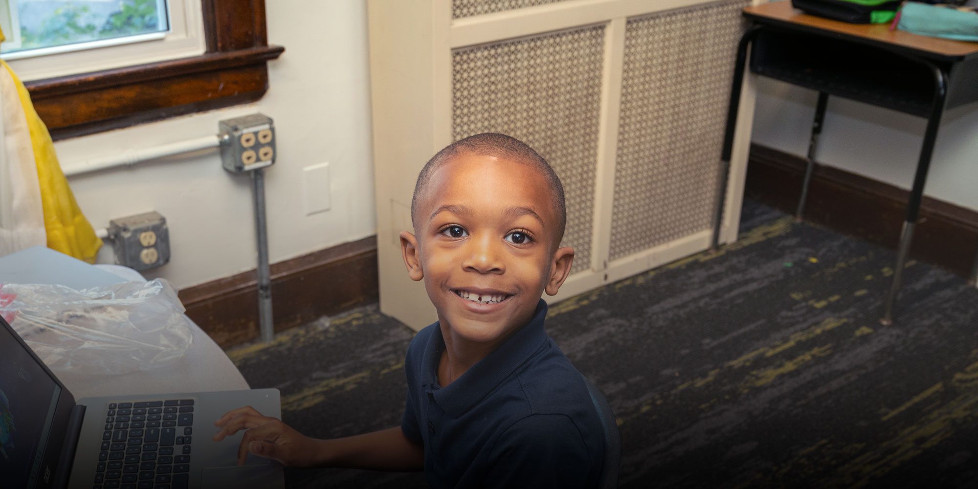 Smiling student looking up from a laptop at a classroom desk.